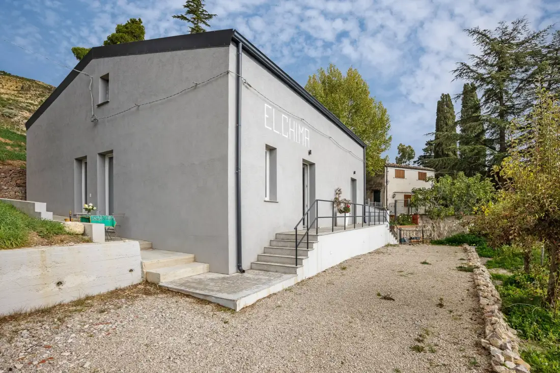 Un edificio gris con escalones exteriores y un patio de grava rodeado de árboles.