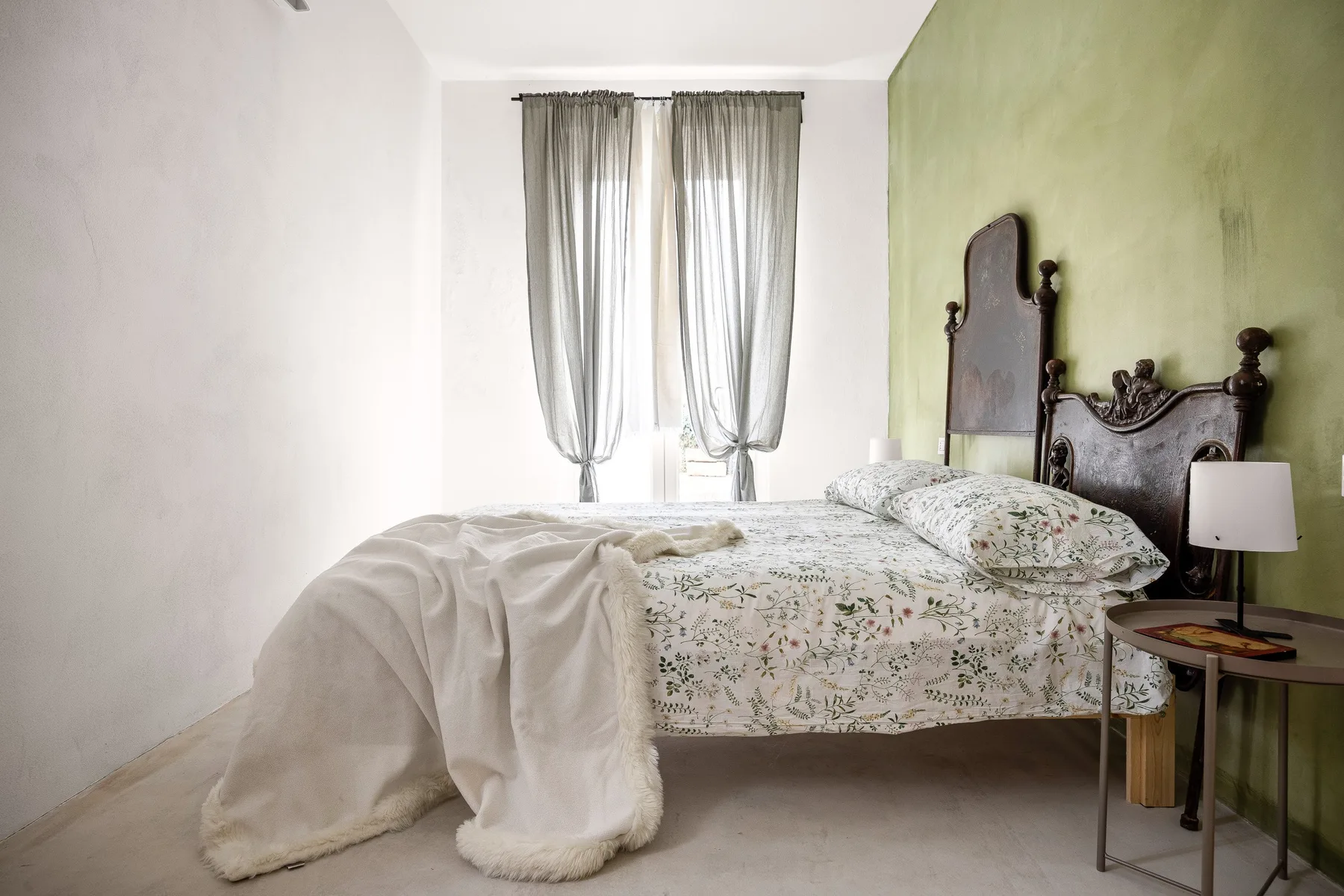 Un dormitorio con una colcha floral, cortinas translúcidas y una mesita de noche.