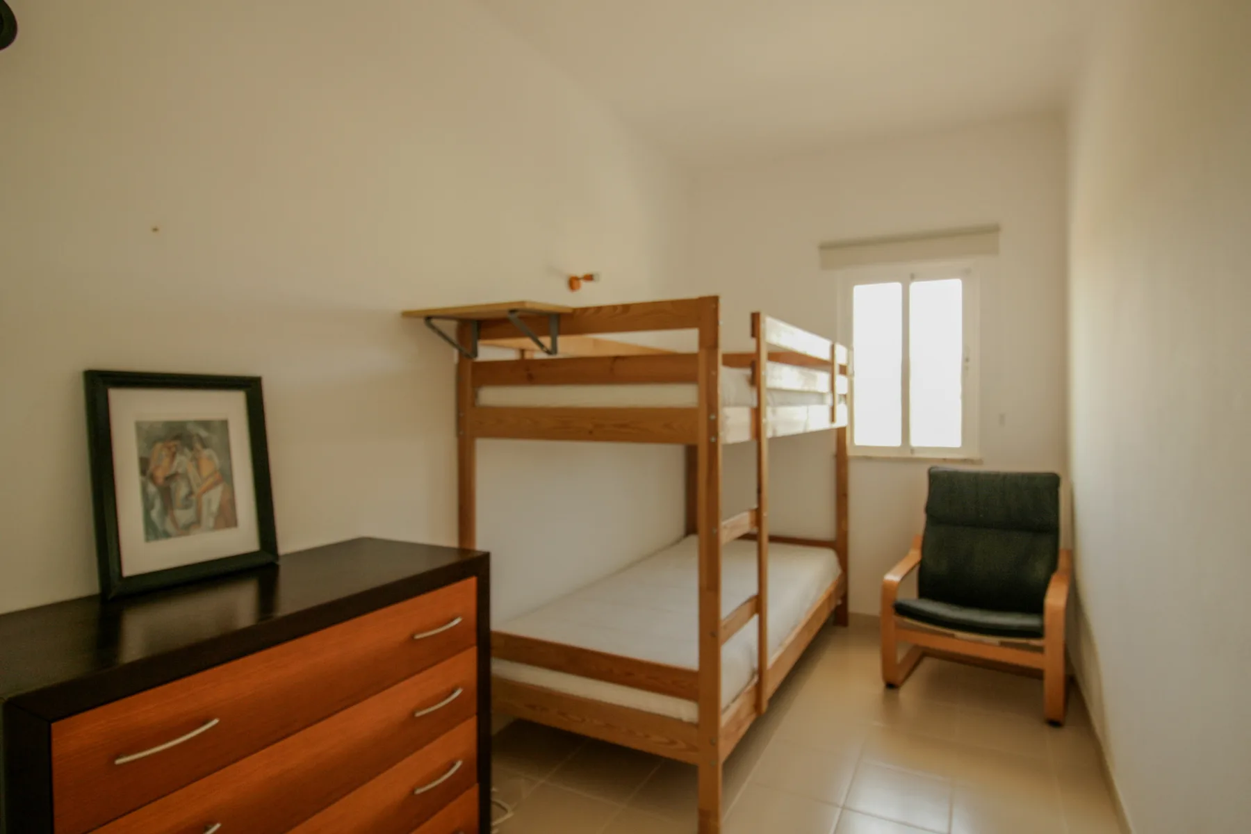 A bedroom with wooden bunk beds, a dresser, and a chair beside the window.