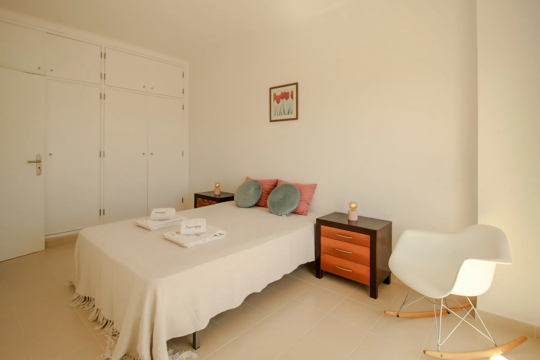 Un dormitorio con una cama tendida, una mesita de noche y asientos blancos.