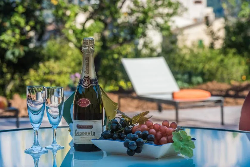 A bottle of sparkling wine, two glasses, and grapes sit on a table beside a pool.