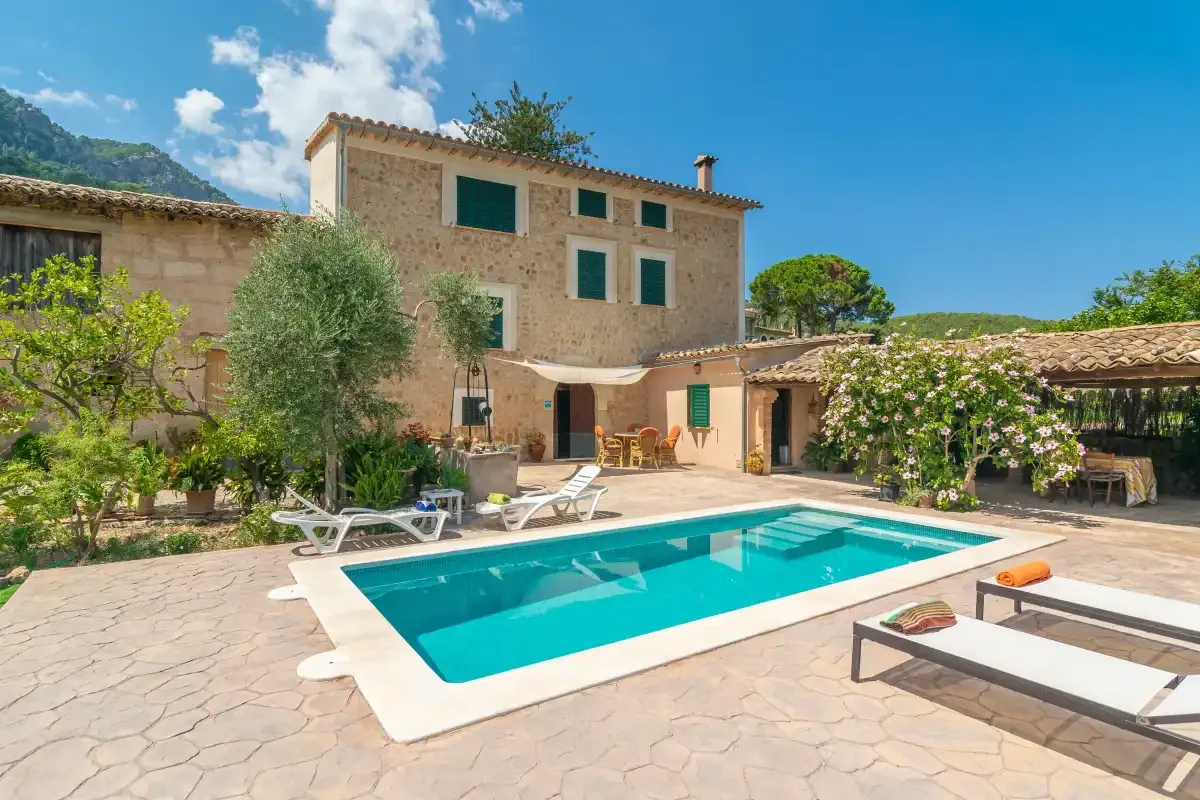 A swimming pool sits in a sunny courtyard beside a stone house.
