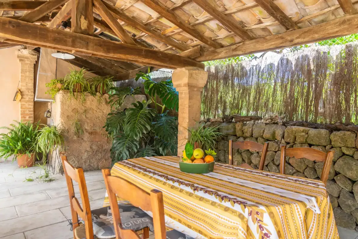A covered outdoor dining area has a table with a patterned cloth and surrounding plants.