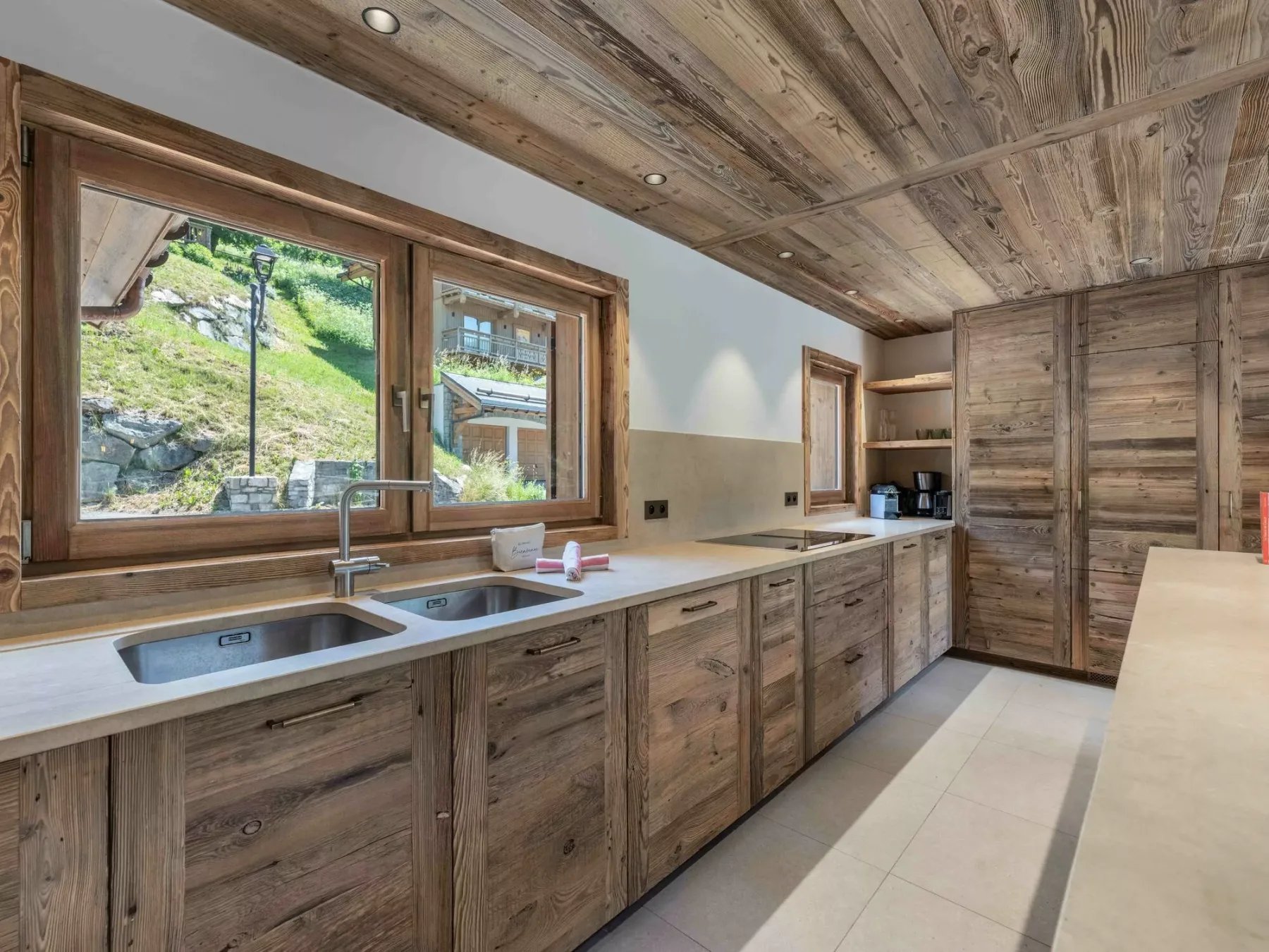 A rustic kitchen with wooden cabinets, a long countertop, and windows overlooking a green hillside.
