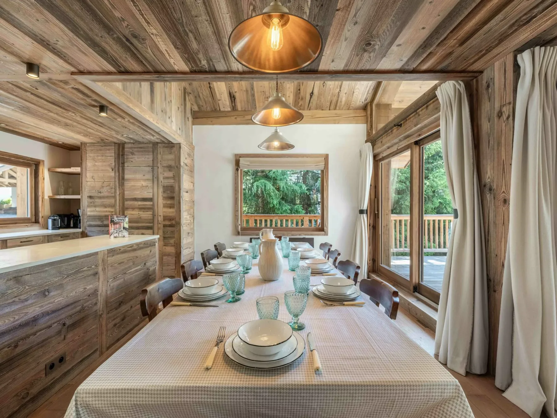 A dining area with a long table set with dishes beneath wooden ceiling beams.