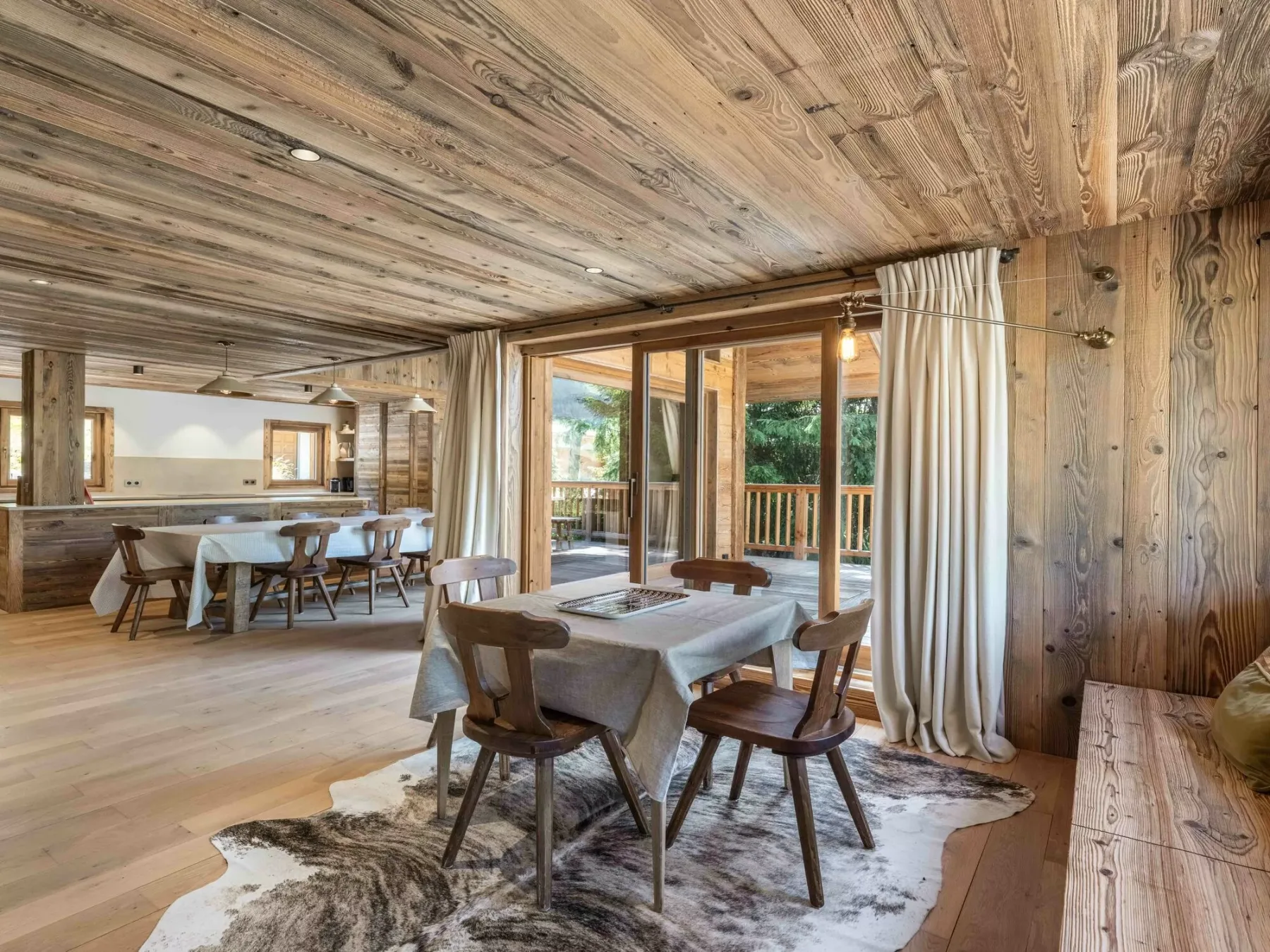 A wooden dining area with tables and chairs beside large windows opening onto a deck.