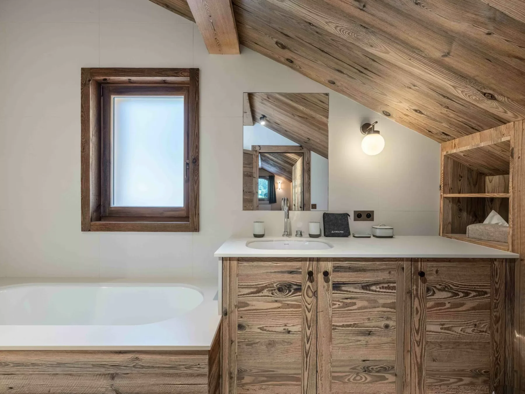 A bathroom vanity and bathtub sit beneath a sloped wood ceiling.