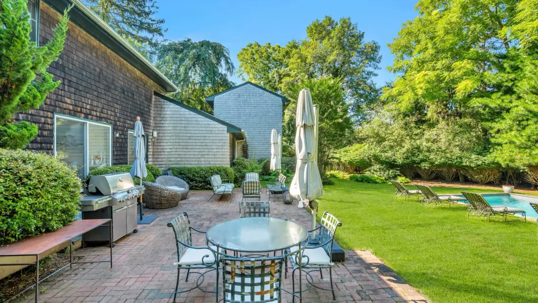 A brick patio with outdoor seating overlooks a lawn and swimming pool.