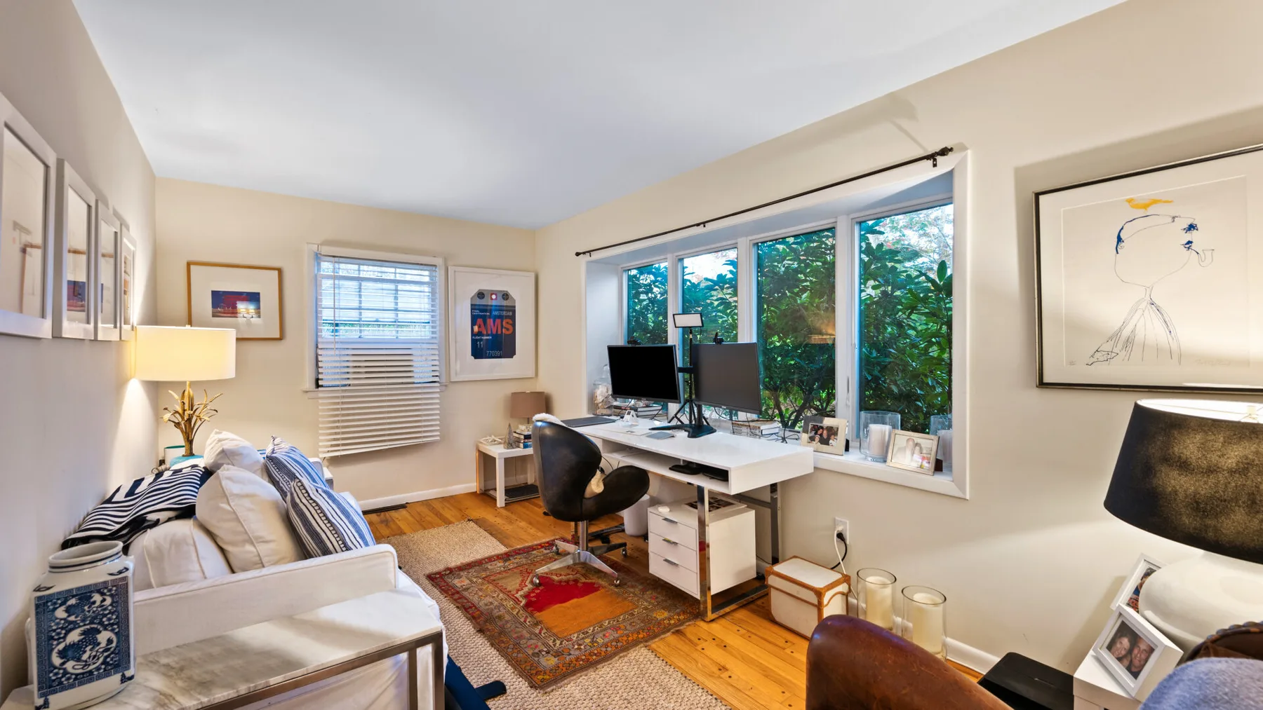 A bright living room includes a seating area beside a desk and large windows.