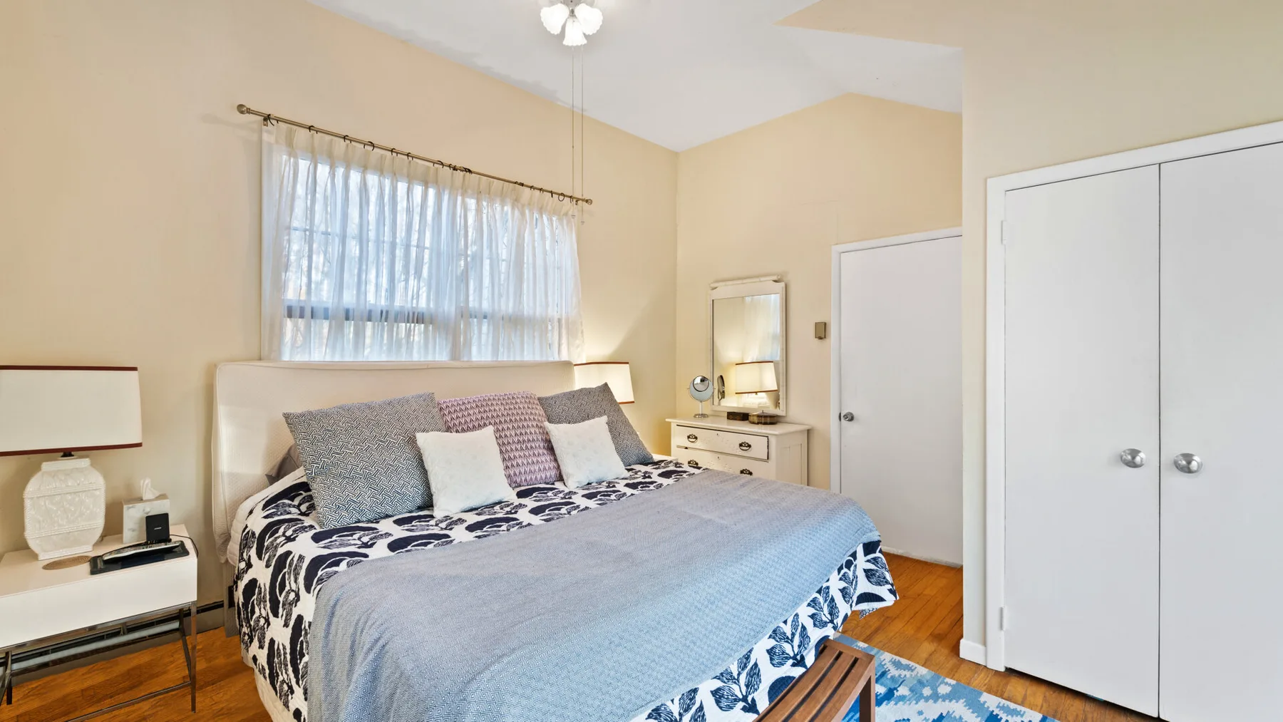 A bedroom with a neatly made bed beneath a window with sheer curtains.