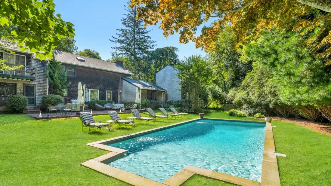 Una piscina está rodeada de césped junto a una casa con árboles.