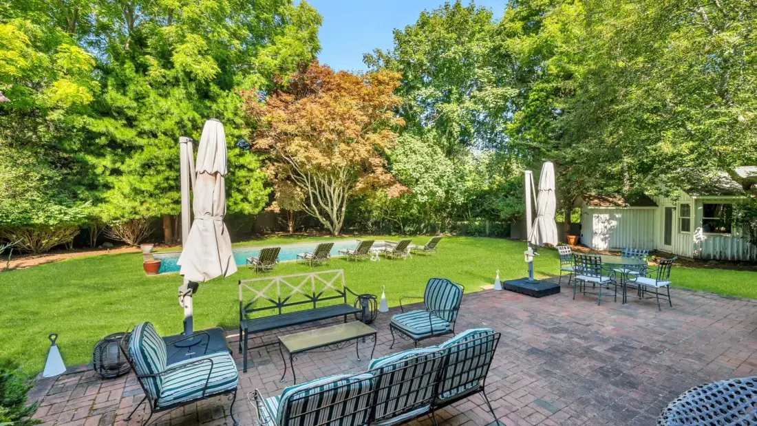 A garden patio with outdoor seating, closed umbrellas, and a lawn surrounded by trees.