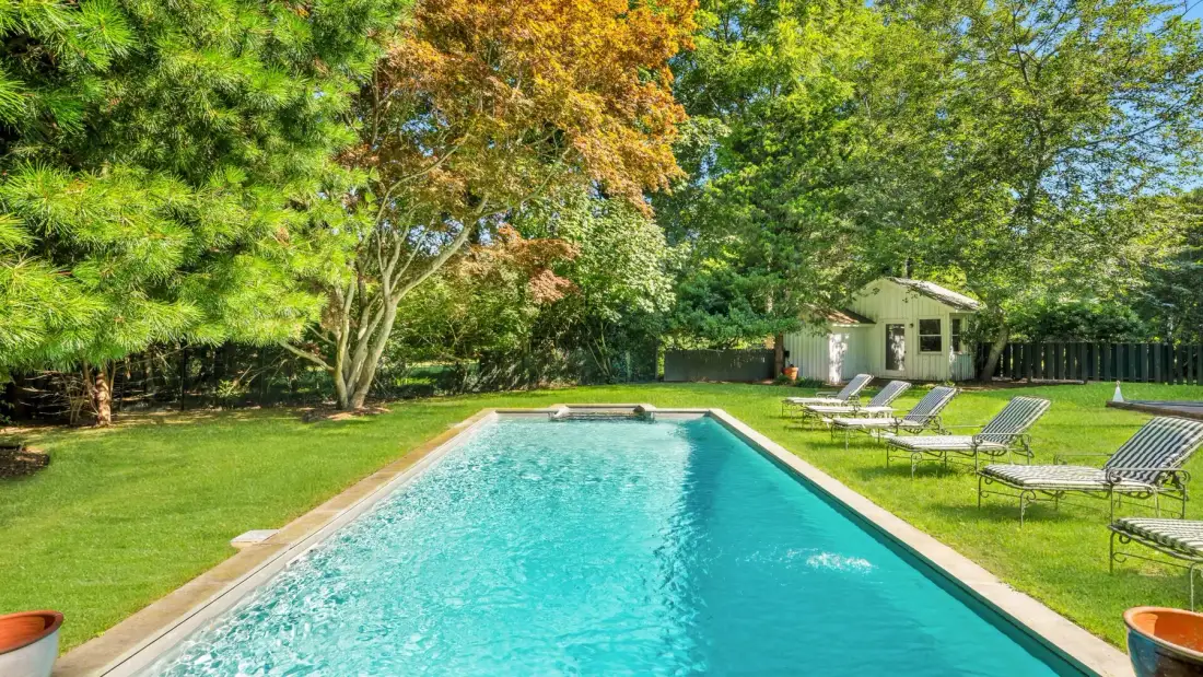 A swimming pool sits in a grassy garden surrounded by trees and lounge chairs.