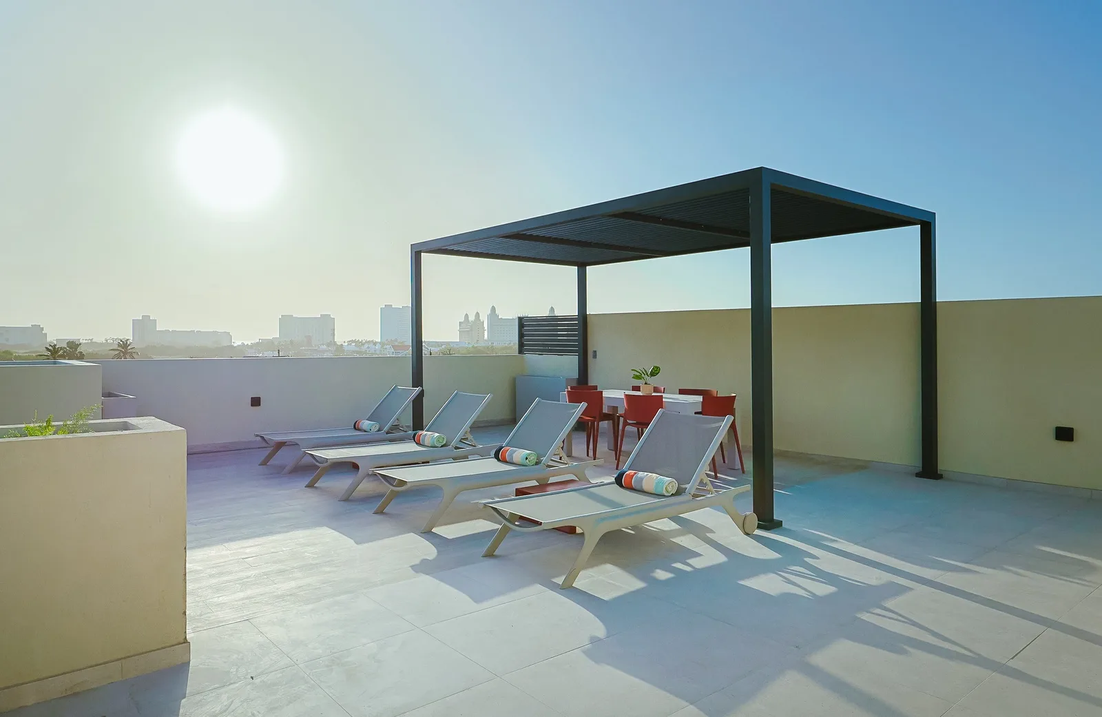 A private rooftop terrace with sun loungers and dining area offers an inviting space under a modern pergola. The urban skyline stretches beyond, setting the stage for bright, sunny days.