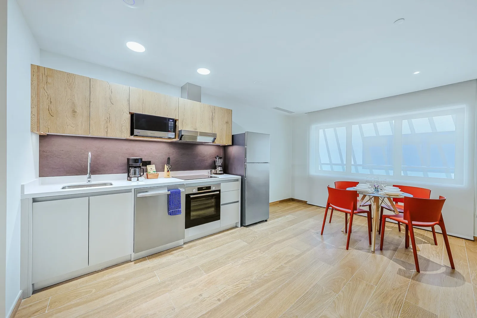 Your fully equipped kitchen and dining area features modern appliances and a bright, open layout enhanced by expansive floor-to-ceiling windows. Bright red dining chairs add a vibrant splash of color, creating a warm and inviting space to enjoy every