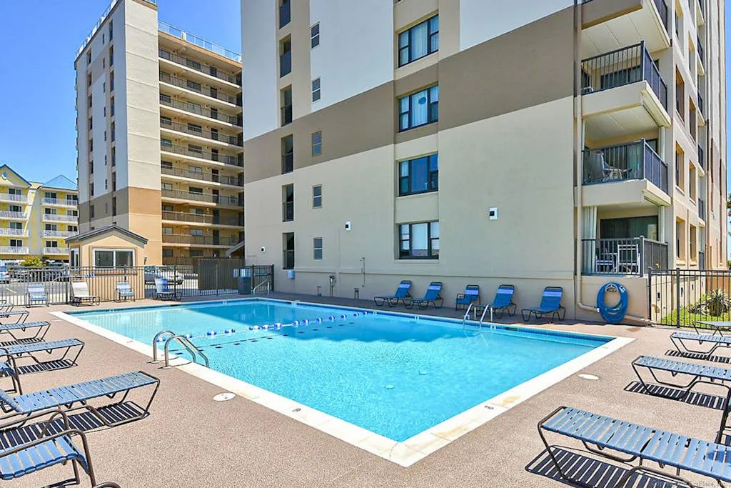 An outdoor swimming pool sits beside a multi-story building, with lounge chairs around the deck.