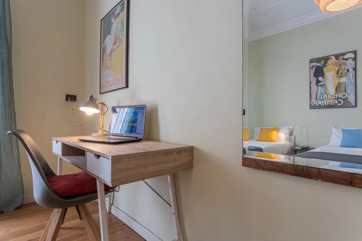 A desk with a lamp and laptop sits beside a mirror reflecting a bed.