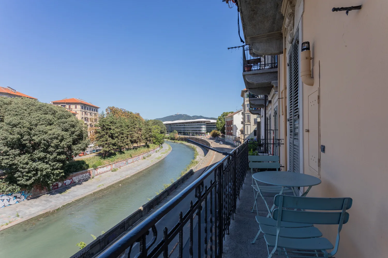 A narrow balcony with seating overlooks a tree-lined canal and nearby buildings.