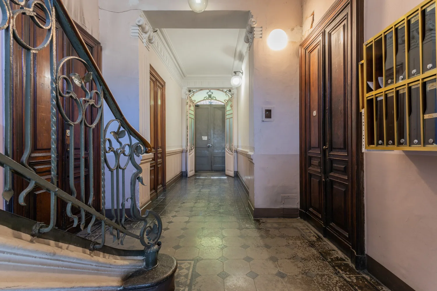 An interior hallway with a tiled floor, ornate staircase railing, and wooden doors.