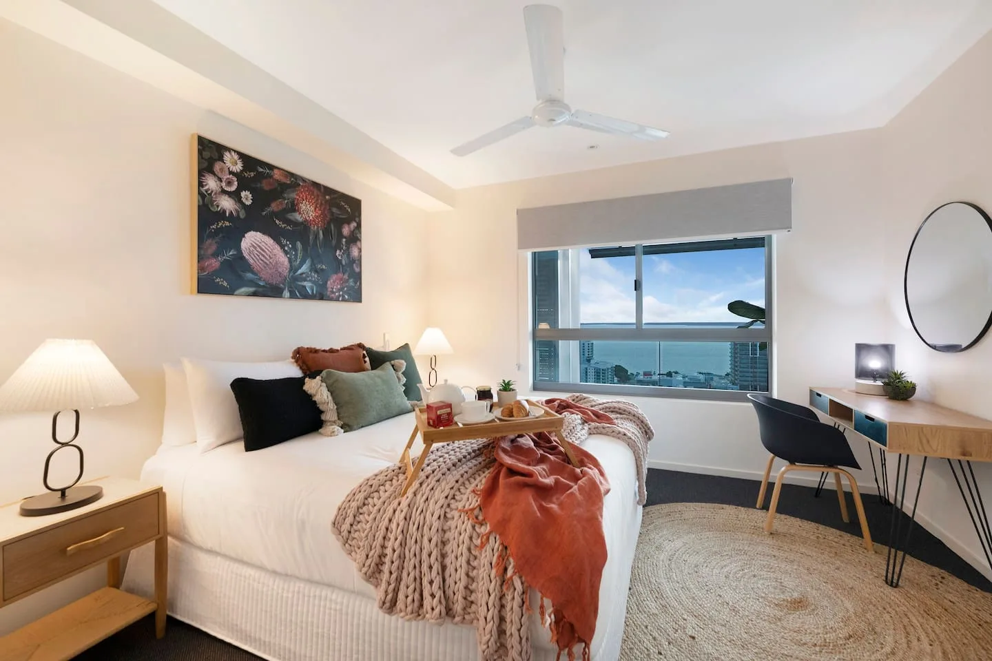 A bright bedroom with a bed, bedside tables, and a desk beside a large window.