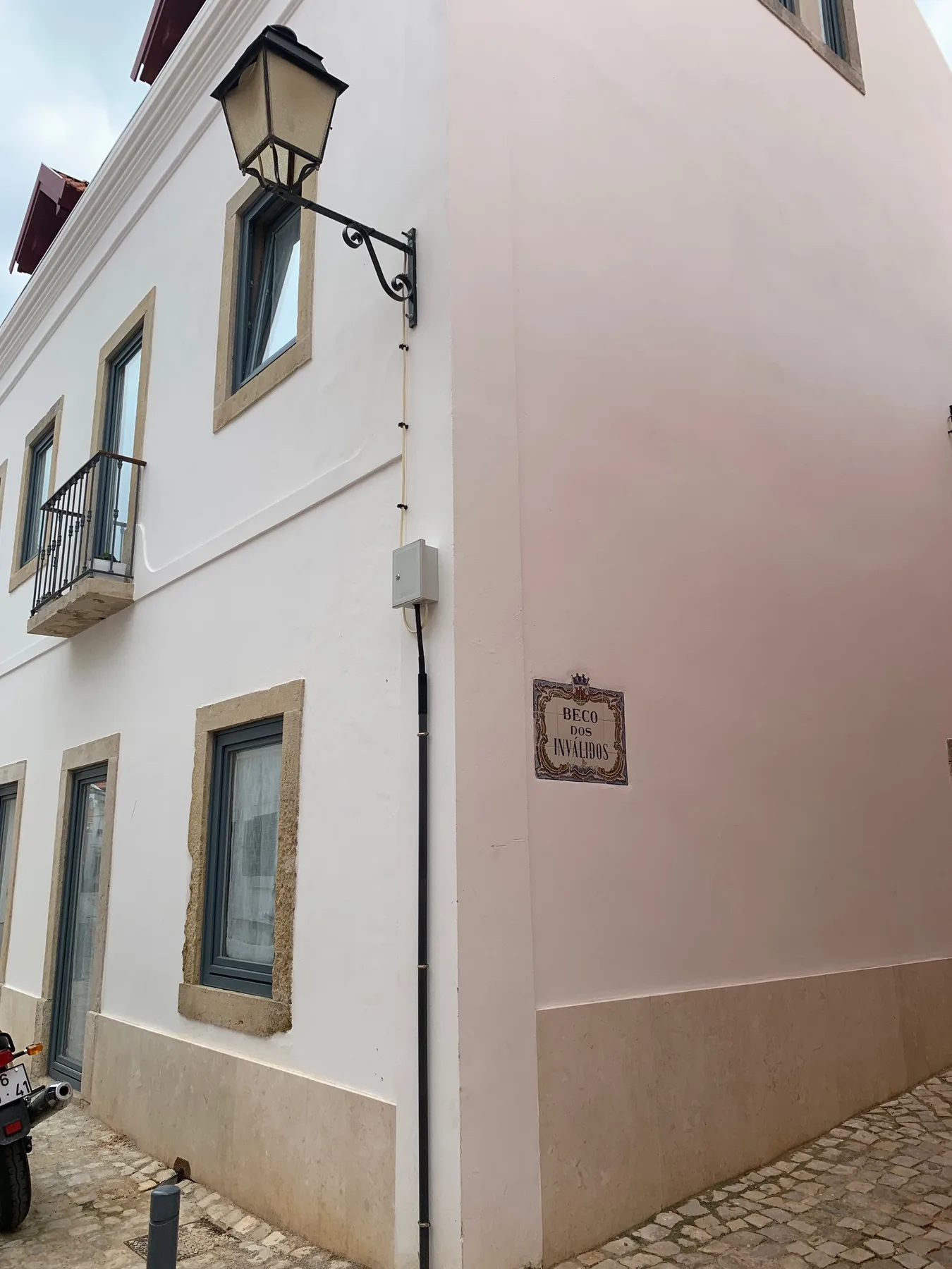 Exterior de un edificio claro con un farol en la pared y pavimento de adoquines.