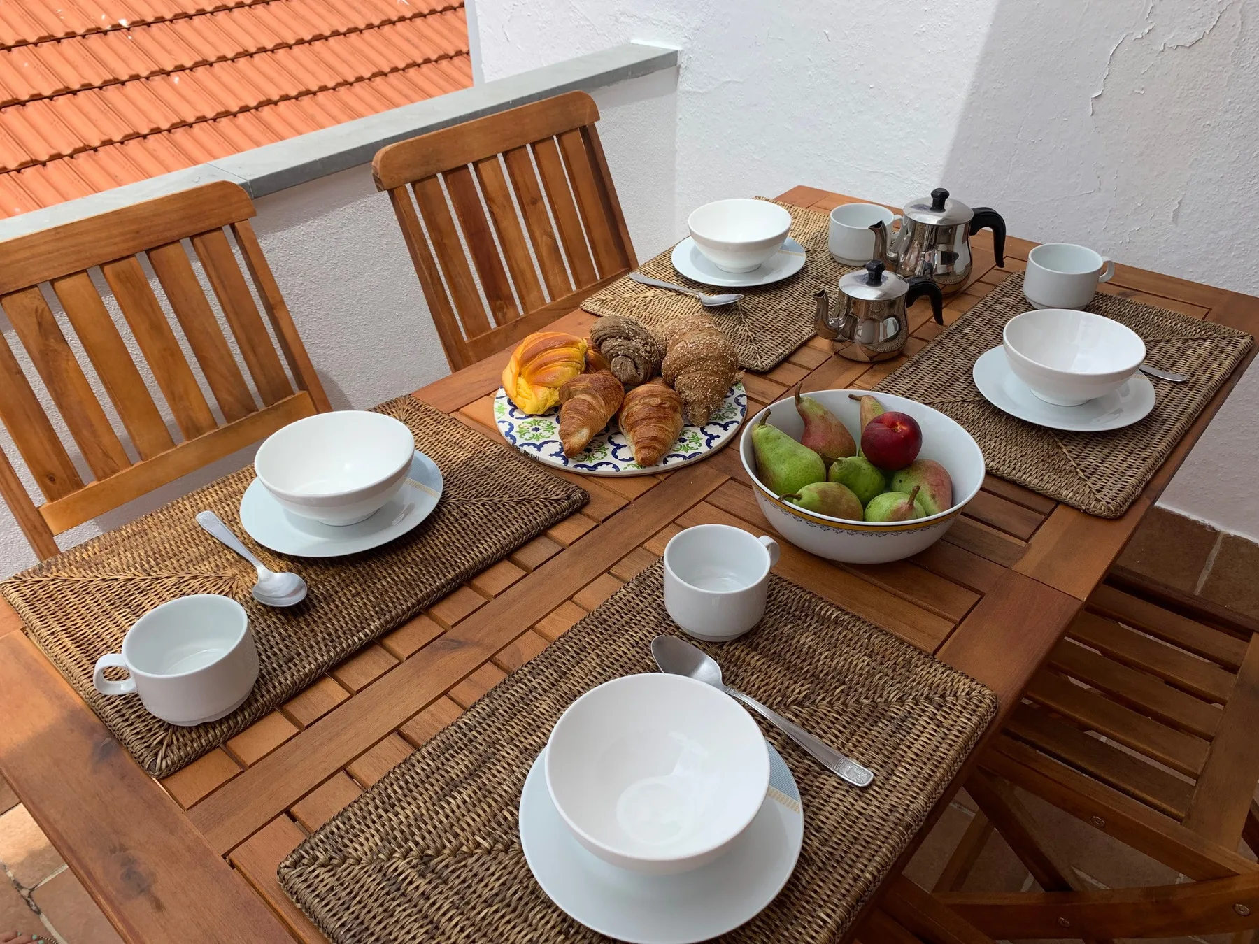 Una mesa de comedor de madera está preparada con platos, tazas y bollería.