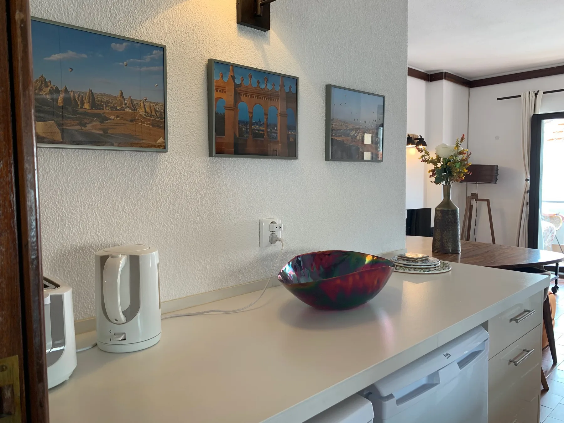 Una encimera de cocina con pequeños electrodomésticos y un cuenco colorido da a un comedor.
