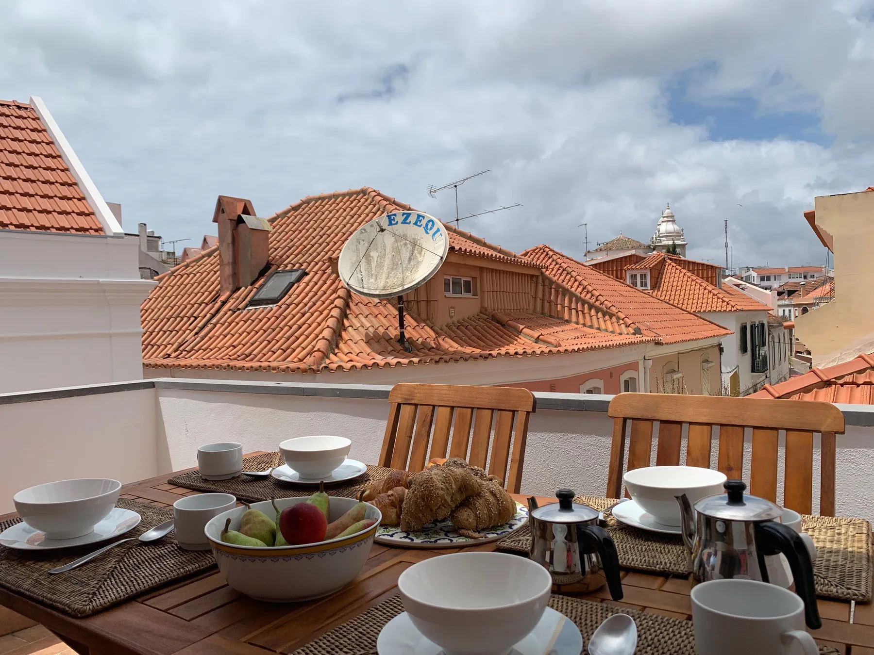 Una zona de comedor en una terraza en la azotea mira hacia tejados de tejas rojas bajo un cielo nublado.