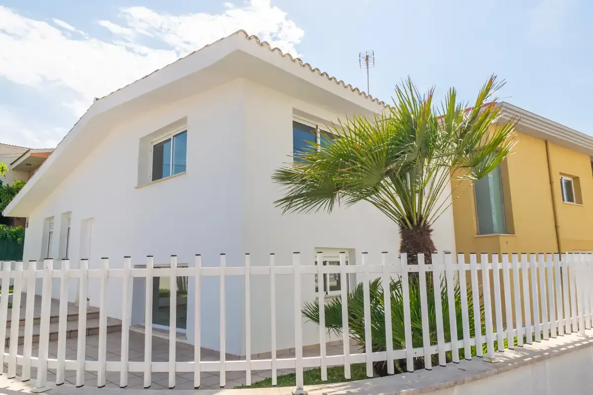Una casa blanca se ve detrás de una cerca blanca, con una palmera delante.