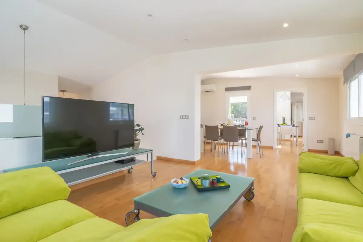Una sala de estar luminosa con asientos verdes y un televisor se abre a un comedor.