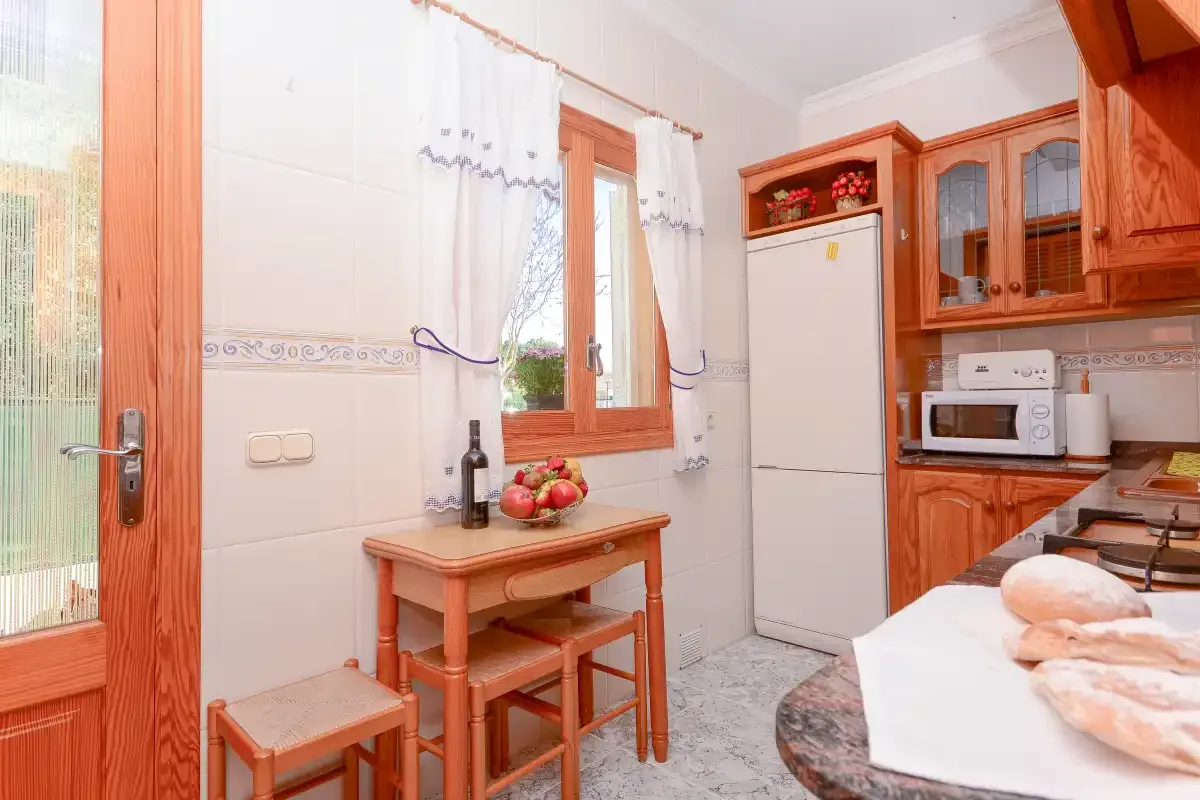 Una cocina luminosa con armarios de madera, un refrigerador y una mesa pequeña junto a la ventana.