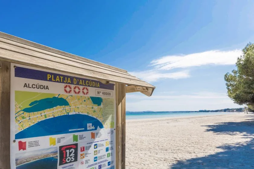 Una playa de arena se extiende junto a un panel informativo de madera bajo un cielo azul despejado.