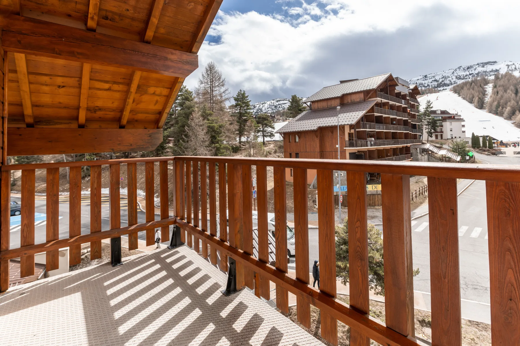 A wooden balcony overlooks snow-covered mountains and nearby buildings.