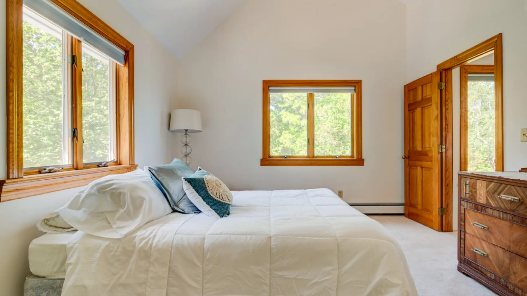 Un dormitorio luminoso con una cama, ventanas con marcos de madera y una puerta de madera abierta.