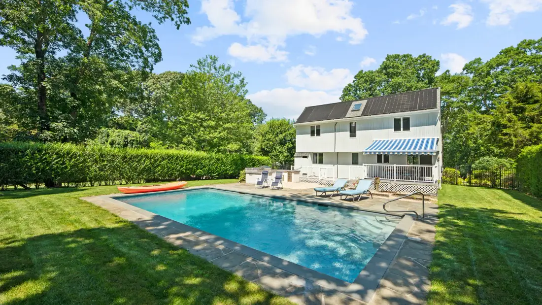 A backyard swimming pool sits in front of a house, with lounge chairs nearby.