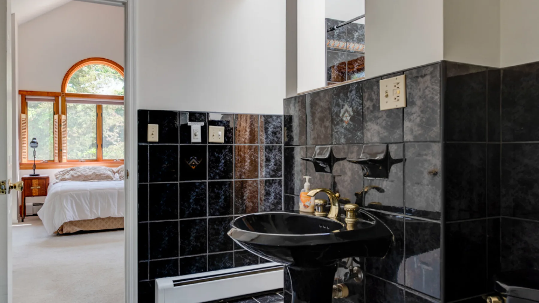Un lavabo de baño con paredes de azulejos oscuros y un dormitorio visible a través de la puerta.