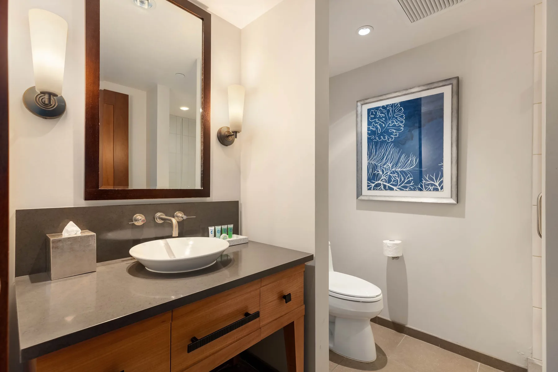 Un lavabo de baño está junto a un inodoro y una obra enmarcada azul.