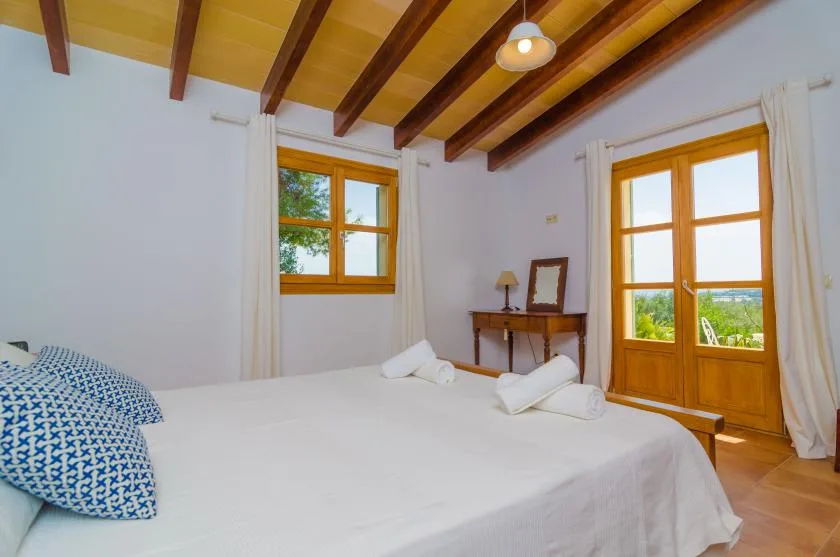 A bright bedroom with a large bed and wooden-framed windows opening onto greenery.