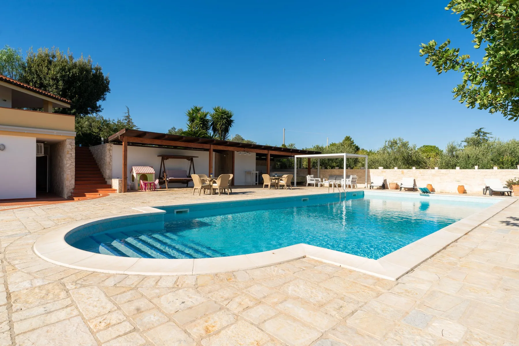 Una piscina está junto a un patio pavimentado con asientos a la sombra.