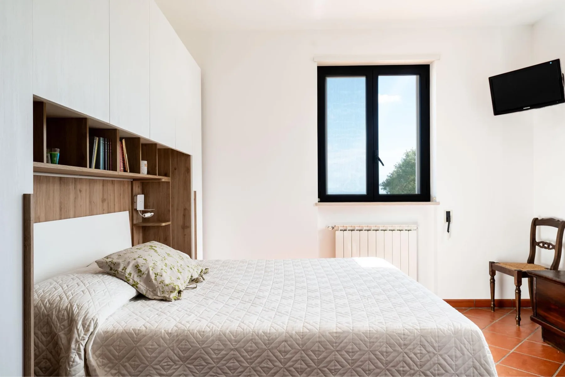 Un dormitorio con una cama grande bajo una ventana y un televisor montado en la pared.