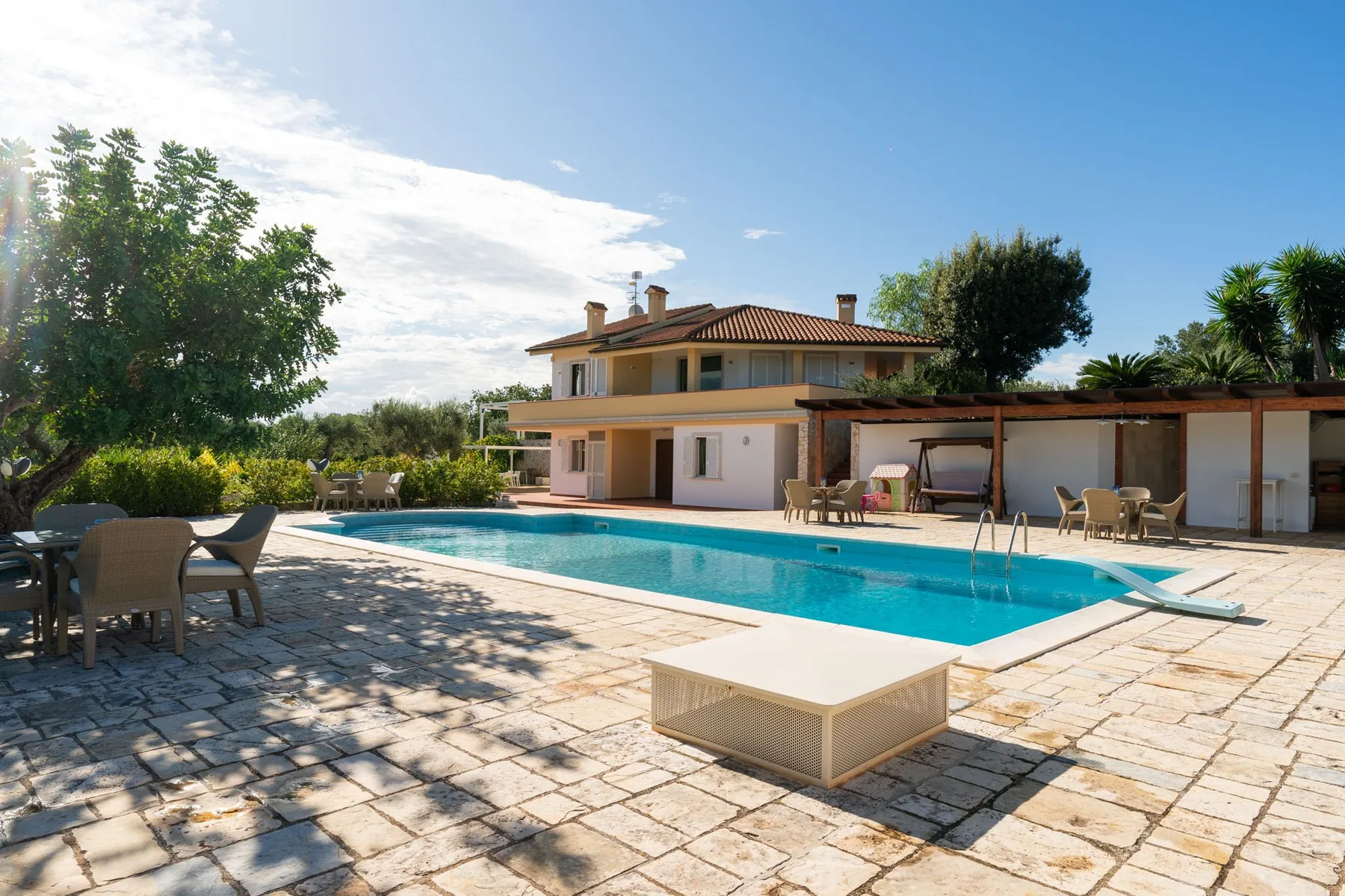 Una piscina está junto a un patio pavimentado con asientos al aire libre y una casa.
