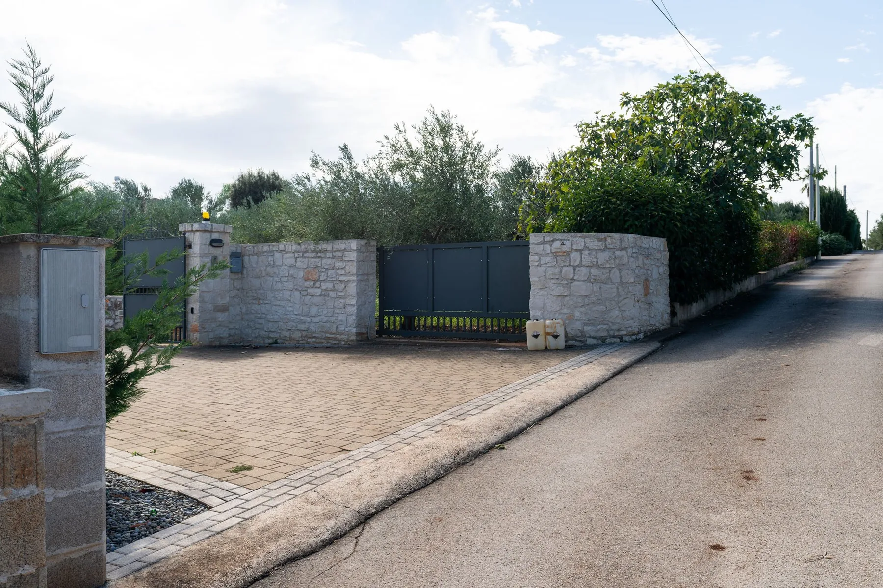 Una entrada pavimentada conduce a un portón oscuro entre muros de piedra, junto a una carretera.