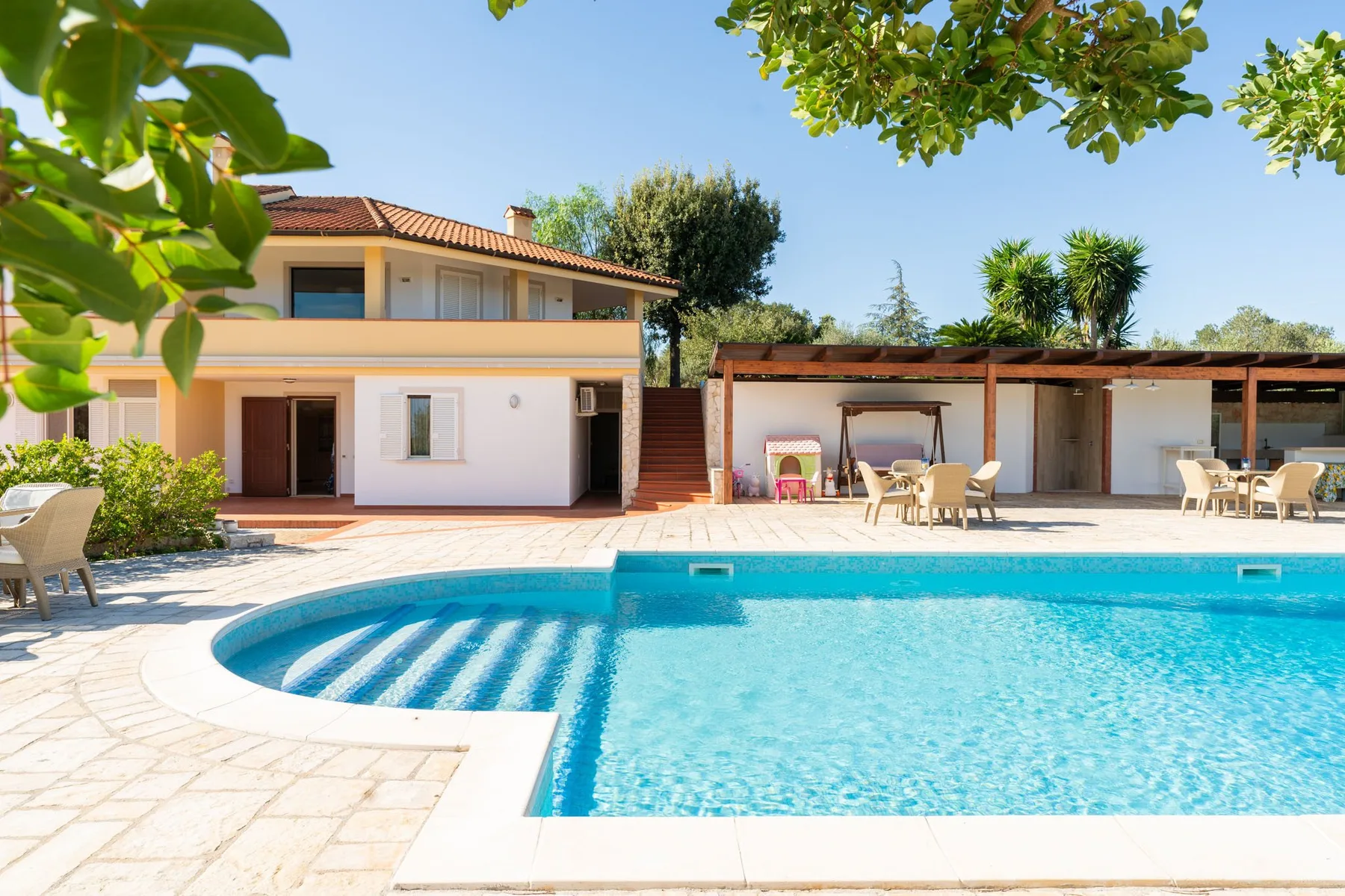 Una piscina está frente a una casa y una zona de estar exterior a la sombra.