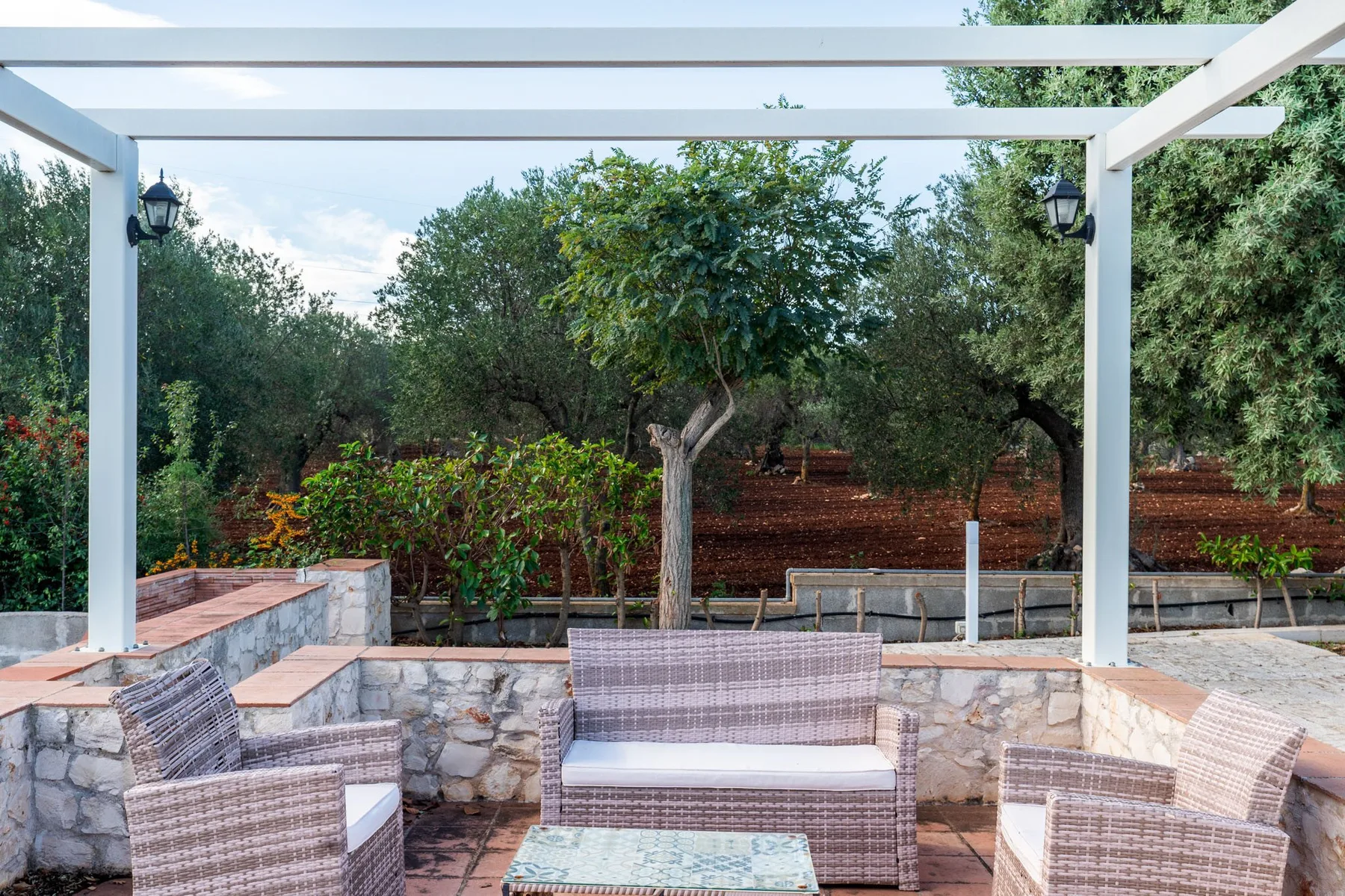 Una zona de estar en un patio bajo una pérgola blanca, frente a un jardín frondoso.