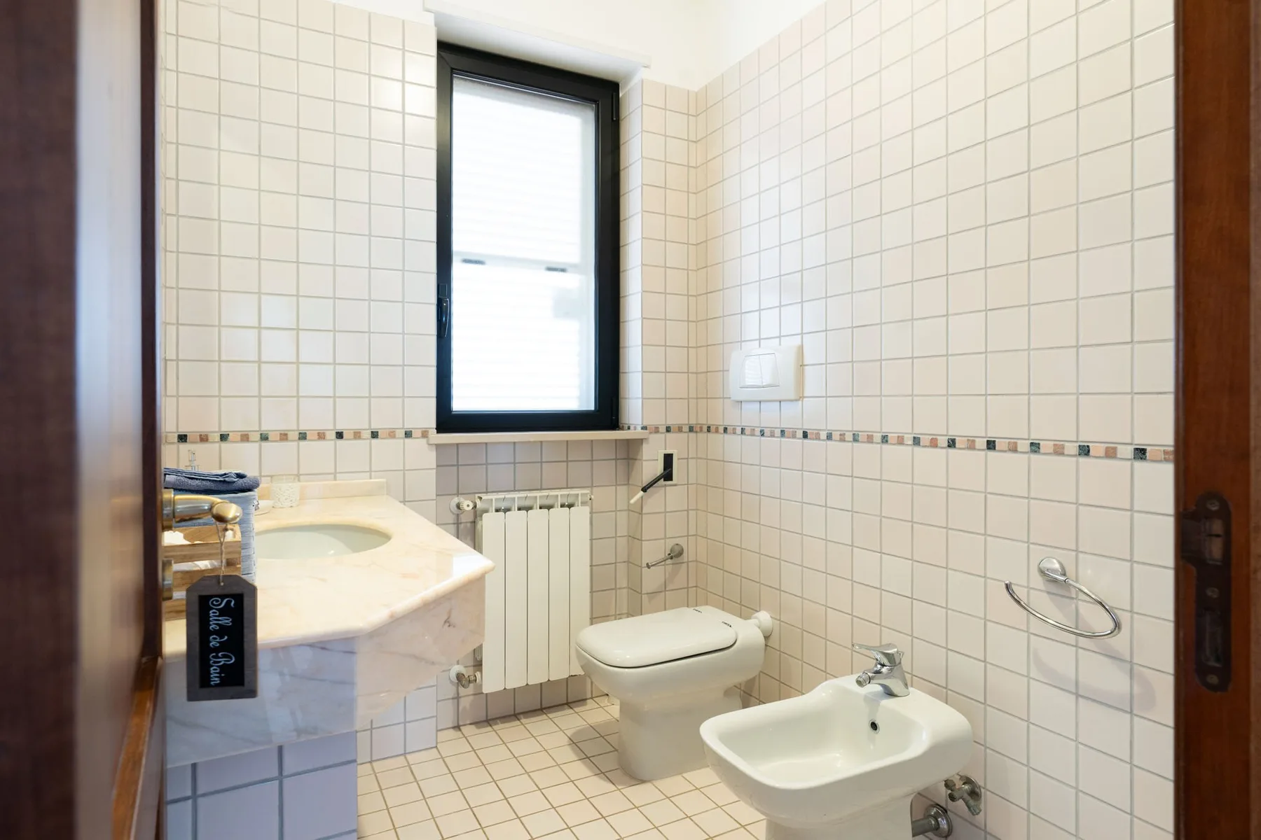 Un baño con azulejos, lavabo, inodoro y bidé bajo una ventana.