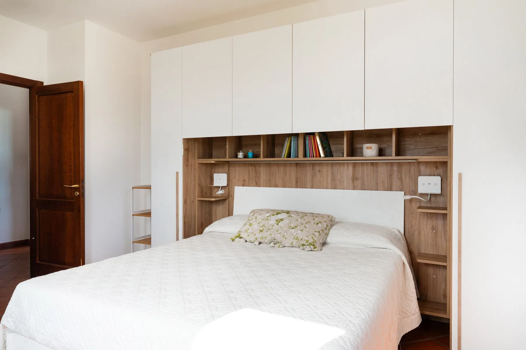 Un dormitorio luminoso con una cama tendida bajo estantes empotrados.