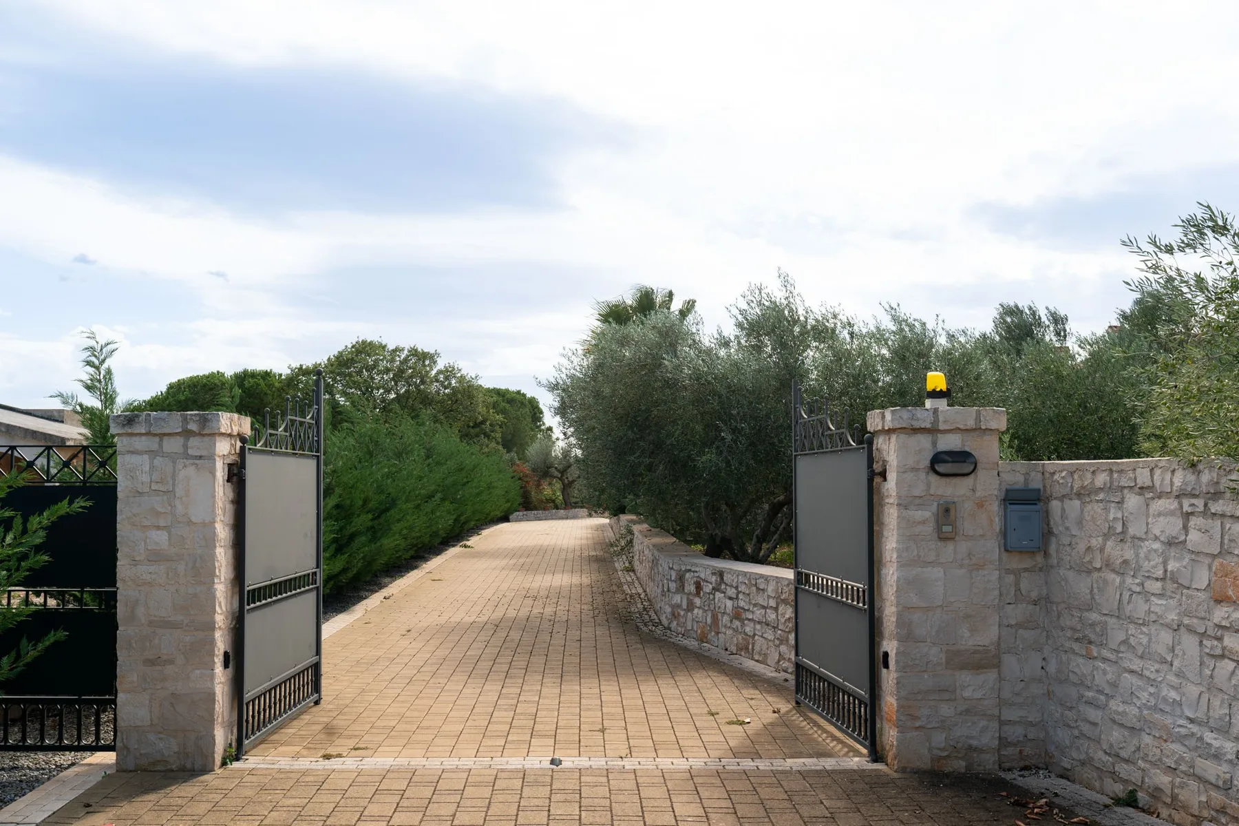 Un camino pavimentado atraviesa una entrada con portones abiertos, árboles y muros de piedra a los lados.