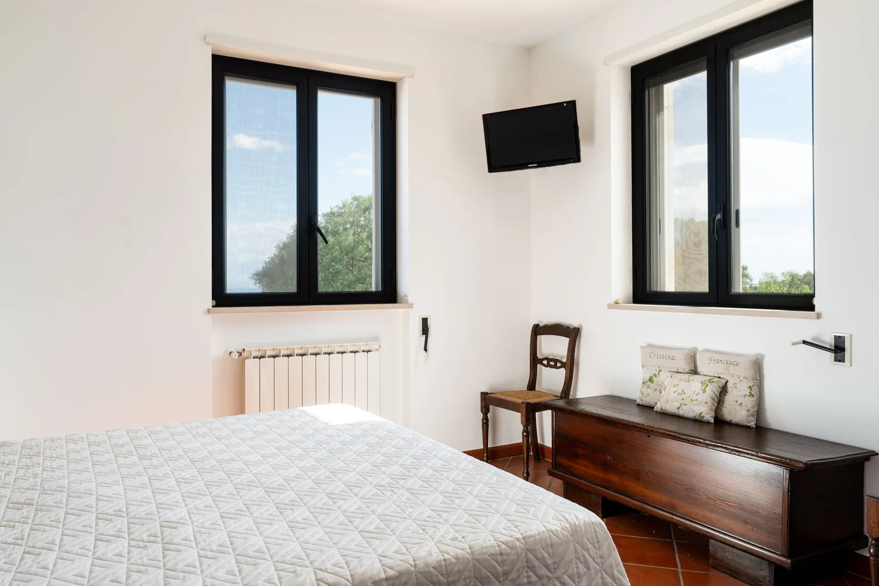 Un dormitorio luminoso con una cama, un banco de madera y dos ventanas grandes.