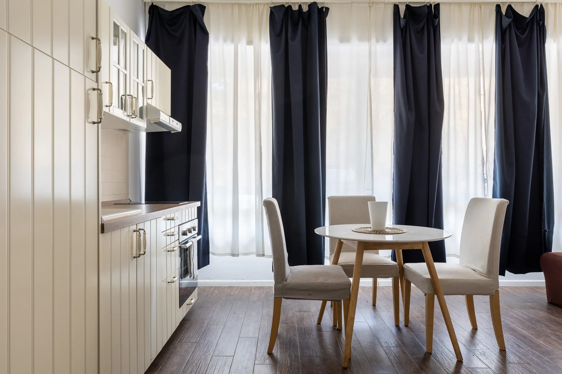 Comedor con una mesa redonda y sillas junto a ventanas altas con cortinas.