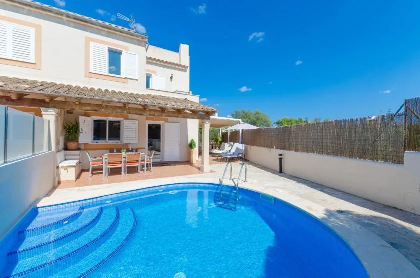 A sunny outdoor pool beside a house, with patio seating under a covered area.